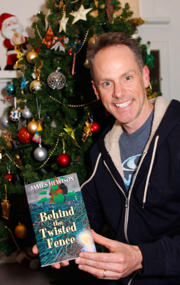 Author James Hewison holds a copy of his children's novel called Behind the Twisted Fence while kneeling in front of a decorated Christmas Tree