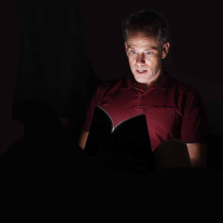 Man in a dark room. Light shines out of a book, lighting up his surprised face.